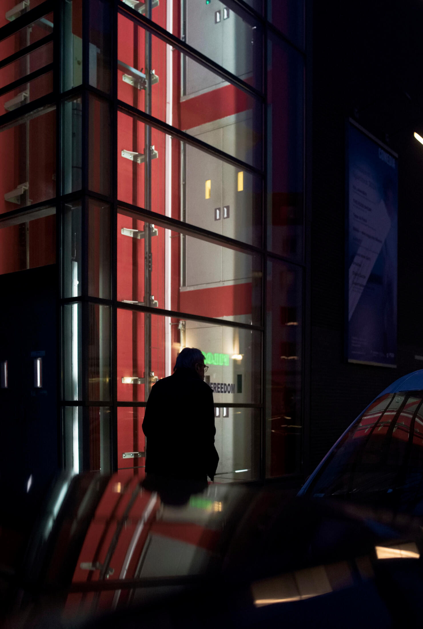 Reflections of Freedom Color street photograph of a solitary figure reflected in illuminated glass architecture at night in Cologne, by Franck Prevot.