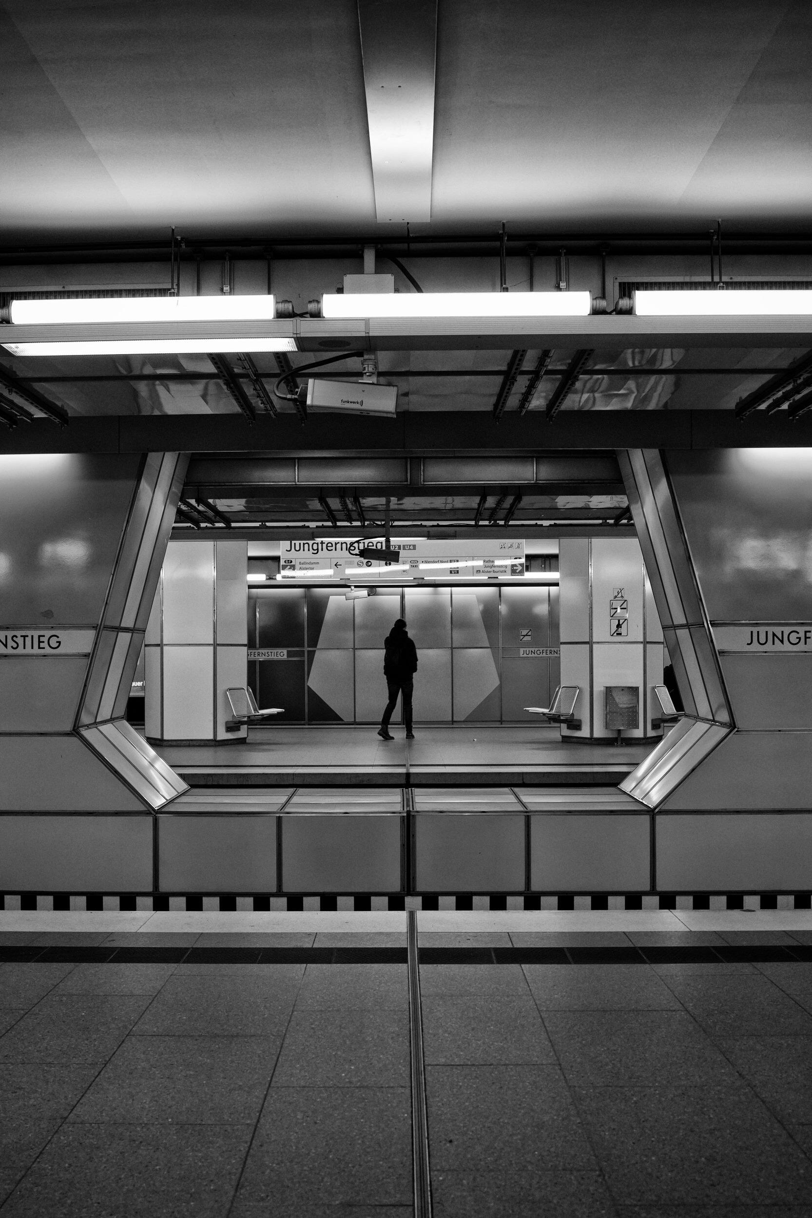 Between Lines Black and white street photograph of a solitary figure in a geometric Hamburg subway station, by Franck Prevot.