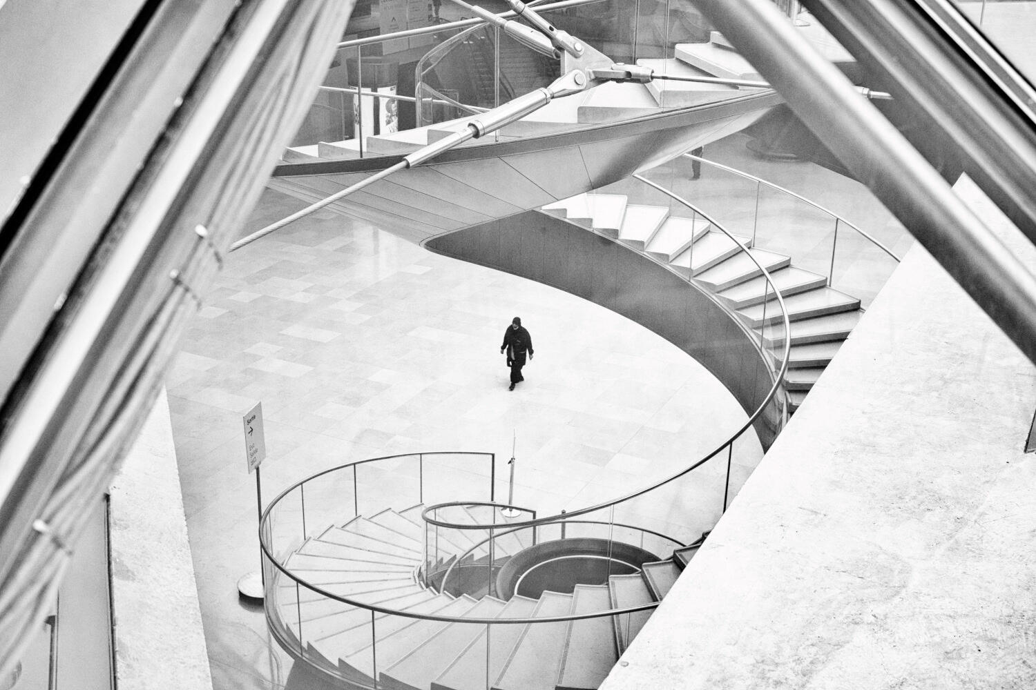 Stillness in Motion Black and white architectural photograph of a solitary figure among curved staircases at the Louvre in Paris, by Franck Prevot.