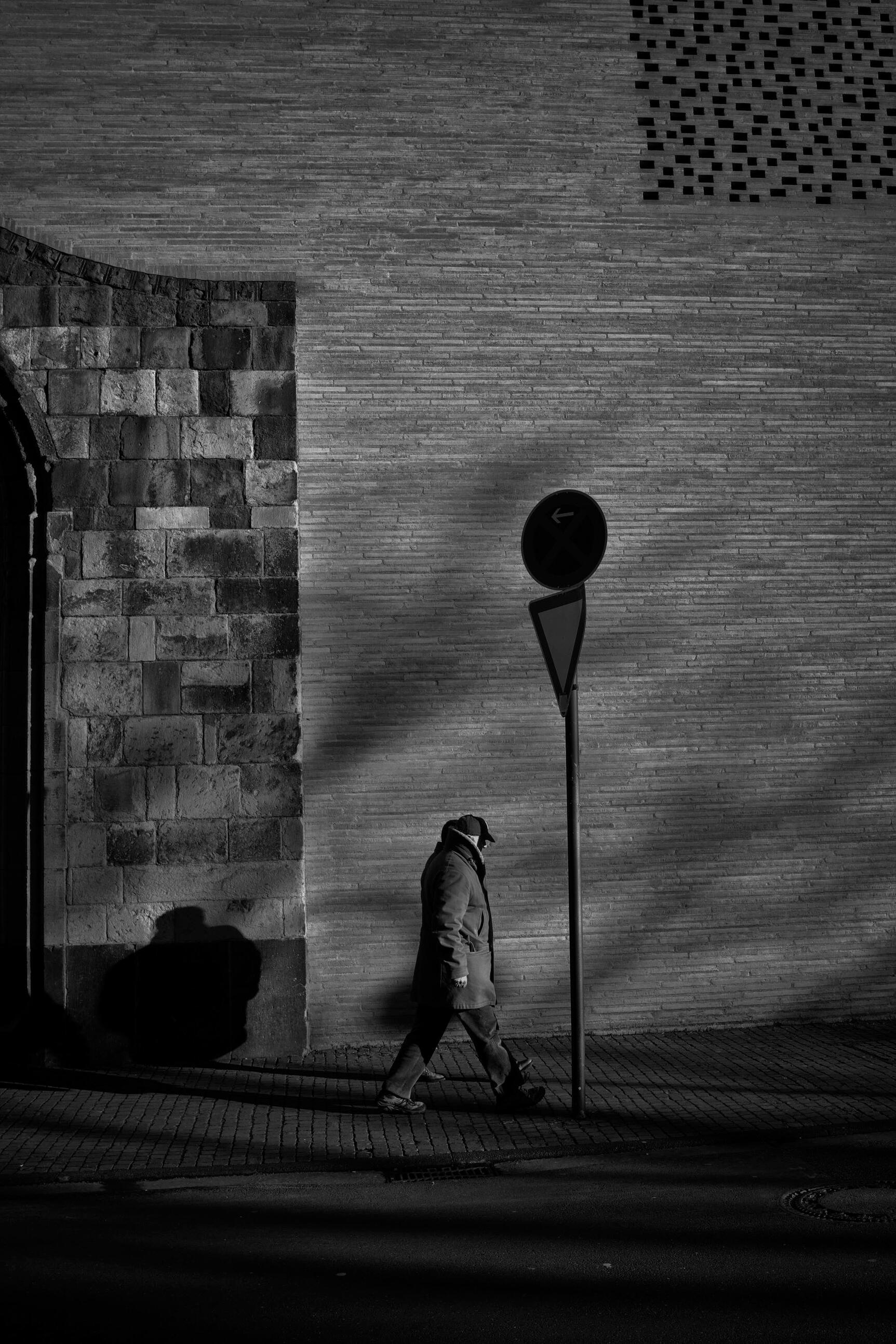 Cologne Noir 8 Black and white street photograph of a lone pedestrian walking through strong light and shadow in Cologne, from the Cologne Noir series, part of the Cologne City Museum collection, by Franck Prevot